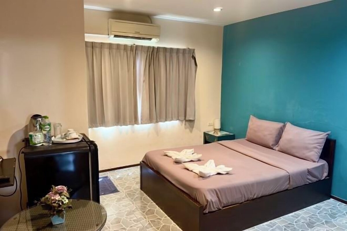 A hotel room with a bed, a bedside table, a chair, a small table with a vase, and a window with curtains.