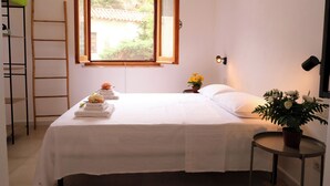A neatly made bed with white linens, a bedside table with a potted plant, a window with a wooden frame, and a wall-mounted light fixture. - Newly refurbished Apartment great location (Buggerru)