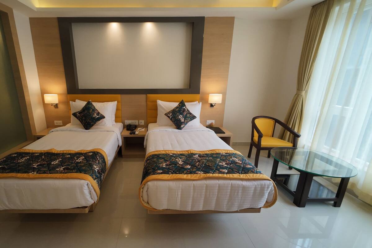 A hotel room with two beds, a glass table, a chair, and a large window with curtains.