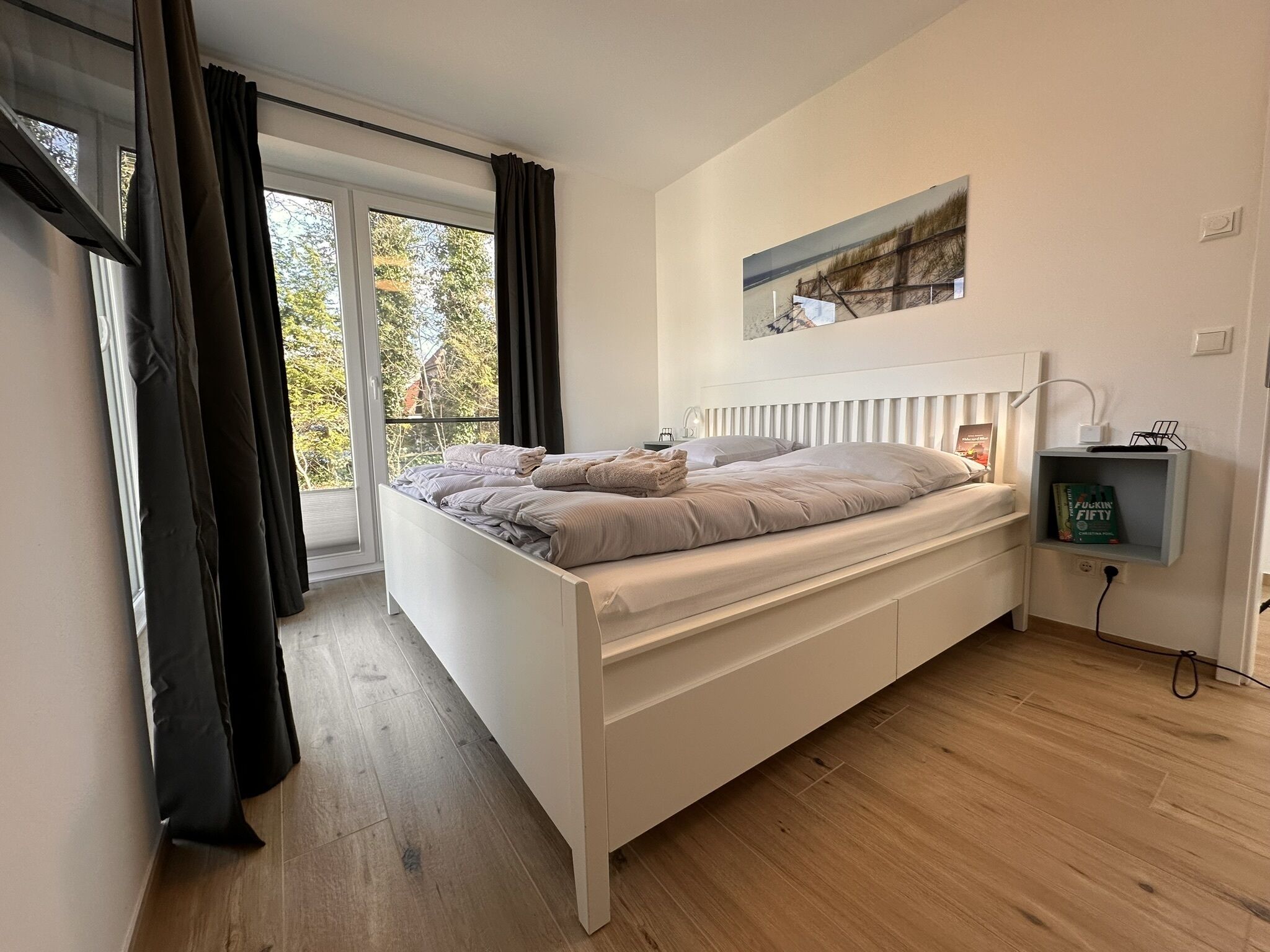 Une chambre moderne avec une grande fenêtre, un lit blanc, une table de chevet avec une lampe et une étagère murale avec un livre.