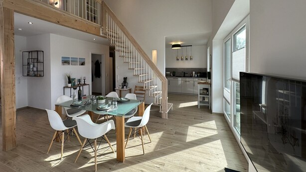 Amplia sala de estar con escalera de madera, una mesa de comedor con sillas y una cocina al fondo.