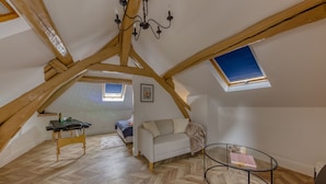A loft bedroom with wooden beams, a slanted ceiling, a small window, a white sofa, a glass table, and a wooden floor.