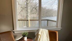A room with a large window overlooking a wooden deck and a body of water. There is a small round table with a plant and a chair with a blanket. - Kayaks | Relax by River | 10 Min to Shipshewana | (Howe)