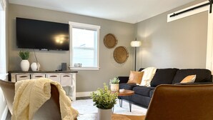 A living room with a sofa, chairs, a coffee table, a TV, a window, and decorative wall items. - Kayaks | Relax by River | 10 Min to Shipshewana | (Howe)