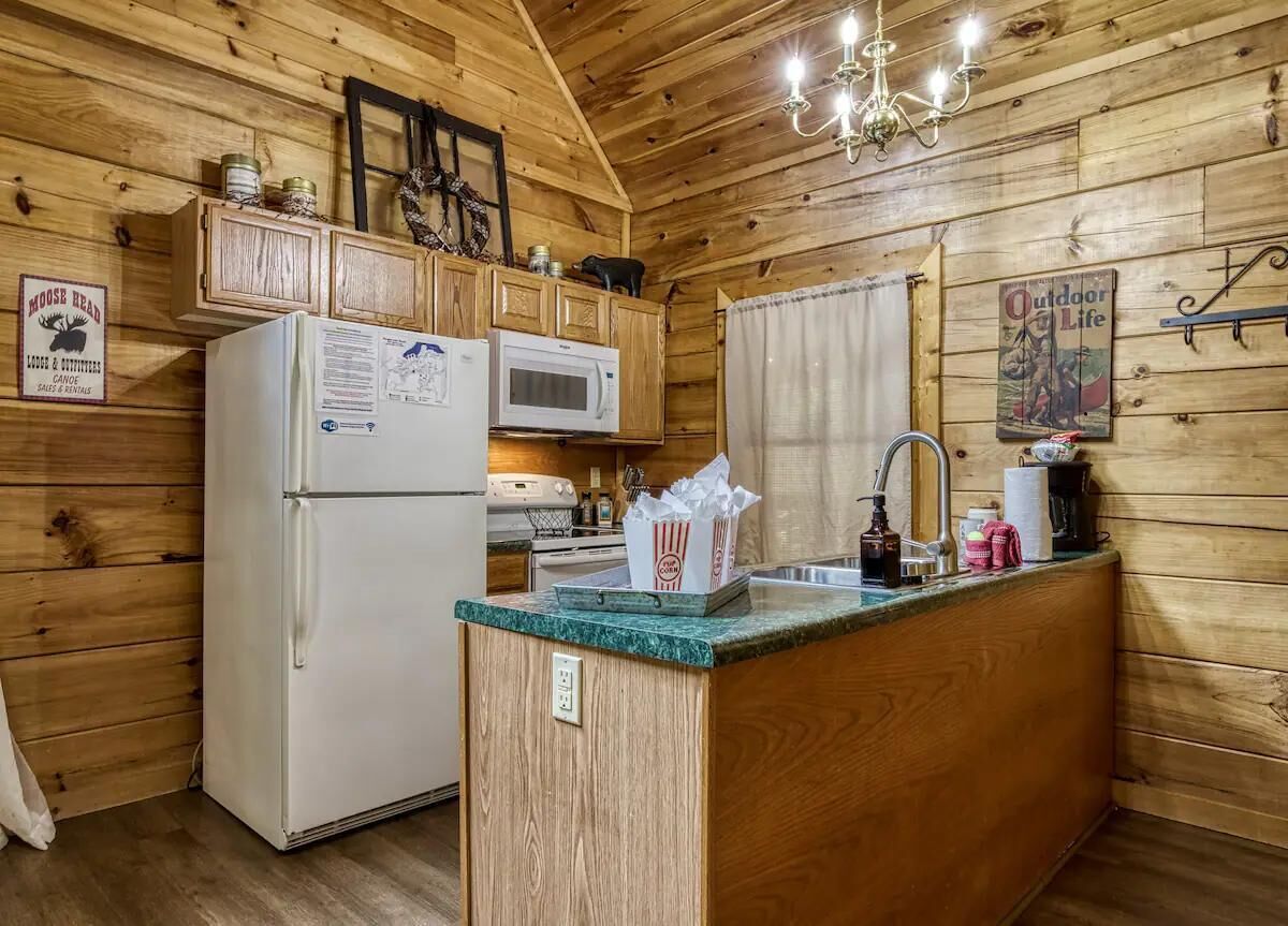 A compact kitchen with wooden walls, a refrigerator, a microwave, a sink, and a chandelier.