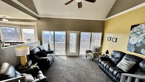 A living room with a ceiling fan, black leather sofas, a lamp, and a large window with a view. - Hummingbird Hideaway (Branson)