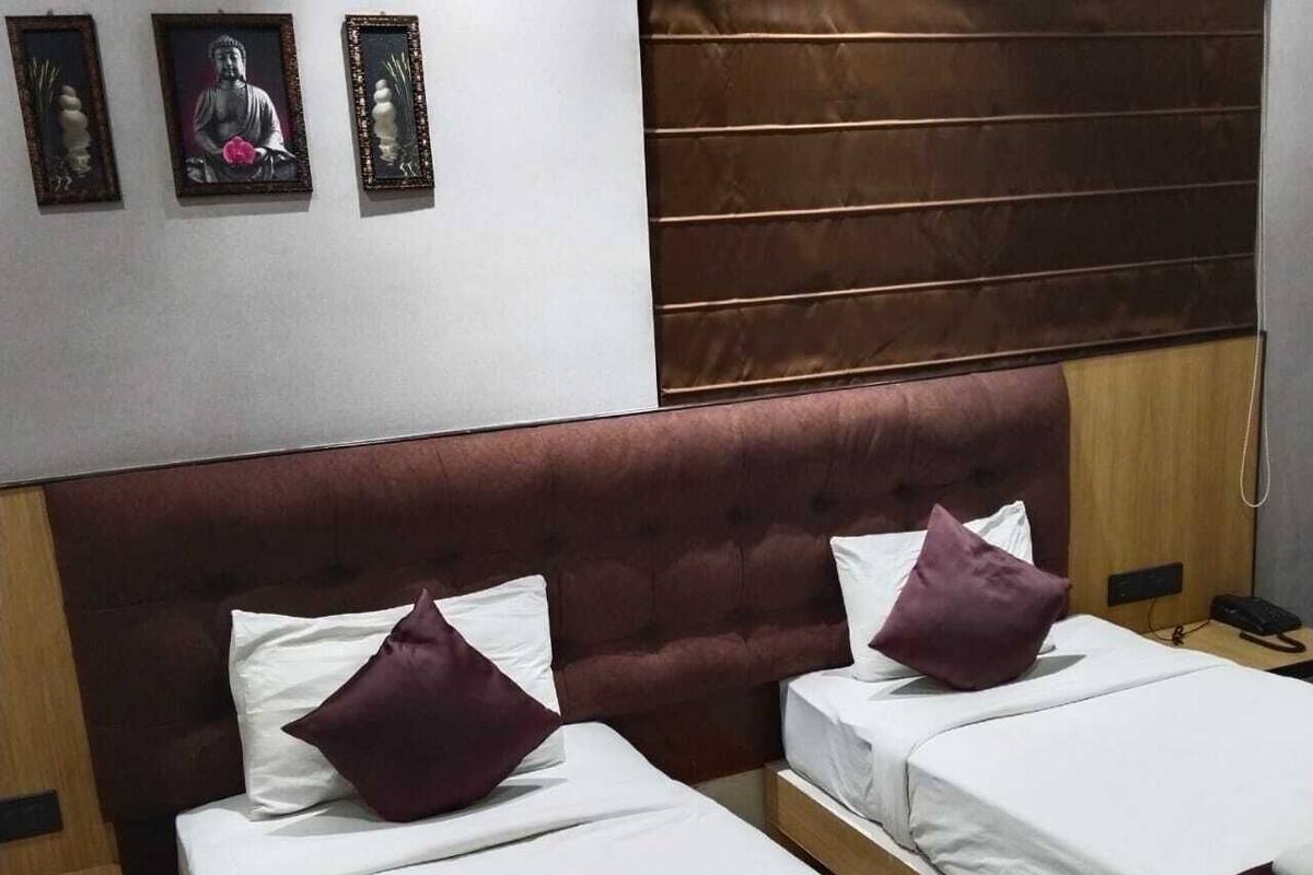 A hotel room with two beds, each with white linens and purple pillows. There is a wooden headboard, a wooden nightstand with a phone, and a wall-mounted shelf with framed pictures.