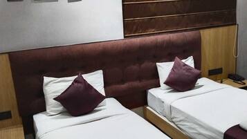 A hotel room with two beds, each with white linens and purple pillows. There is a wooden headboard, a wooden nightstand with a phone, and a wall-mounted shelf with framed pictures.