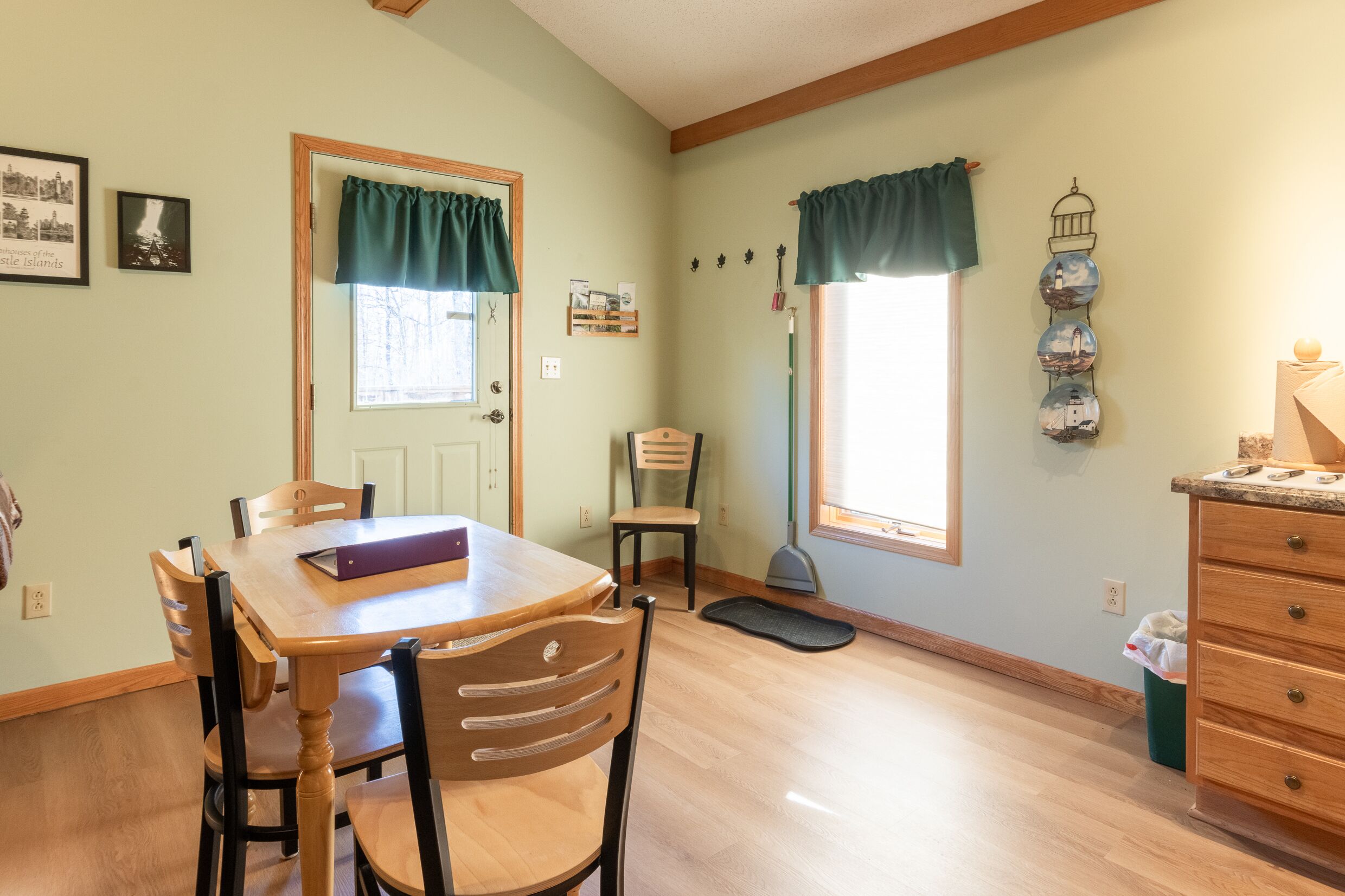 A dining area with a wooden table and chairs, a window with curtains, a broom, and a wall-mounted decoration.