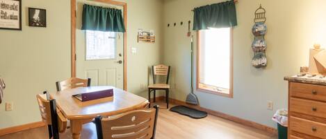 A dining area with a wooden table and chairs, a window with curtains, a broom, and a wall-mounted decoration.