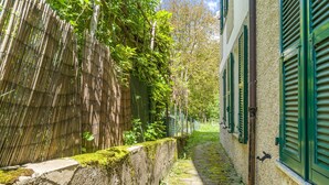 Un étroit sentier pavé, bordé d’une clôture en bambou et d’un mur de pierre, mène à un bâtiment aux volets verts.