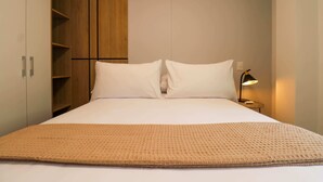 A neatly made bed with white linens, a beige throw blanket, and a wooden wardrobe with multiple shelves.