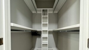 Un vestidor estrecho con paredes blancas, un estante central con escalera y suelo de madera gris.