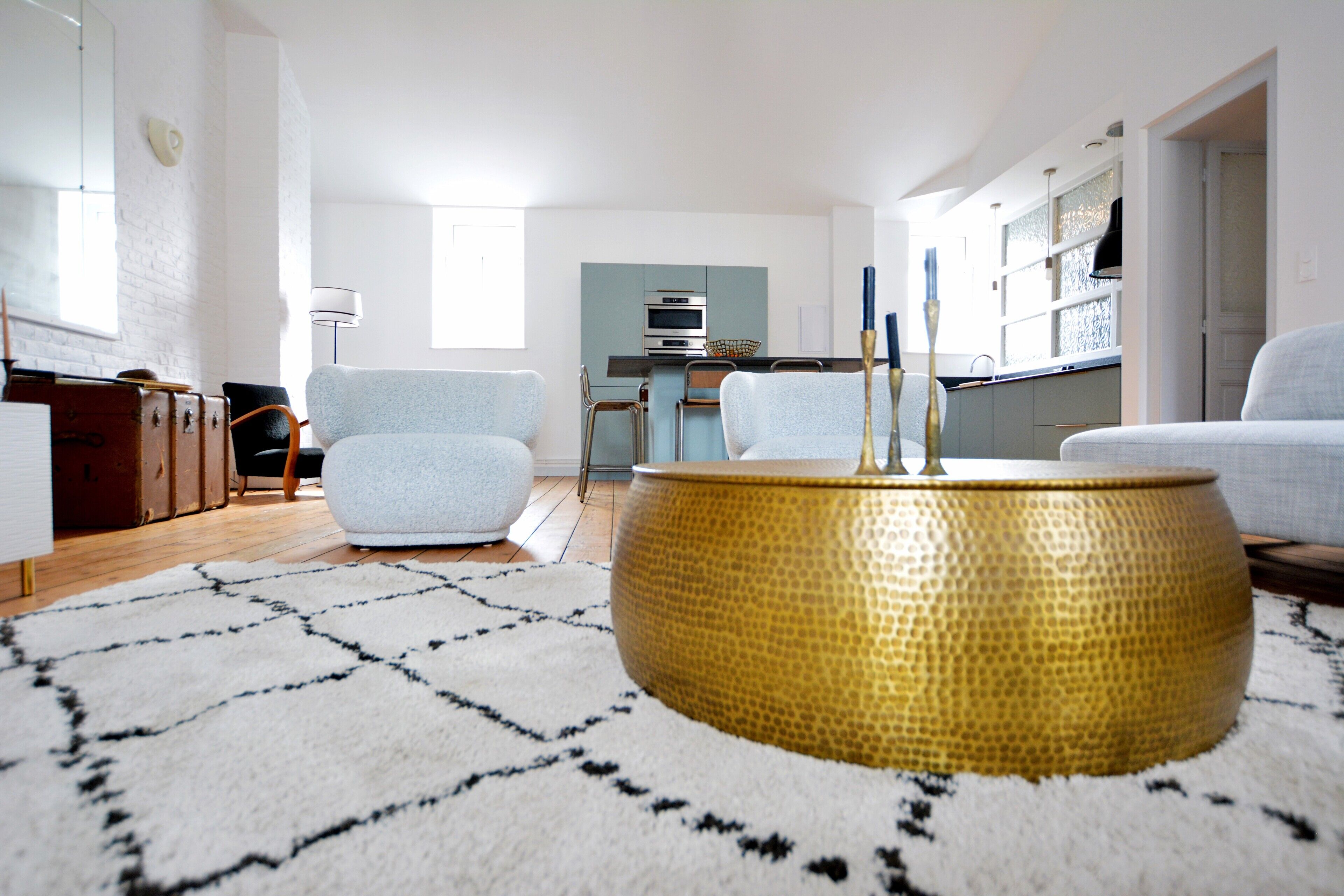 A modern living room with a round gold table, light blue armchairs, and a kitchen in the background.