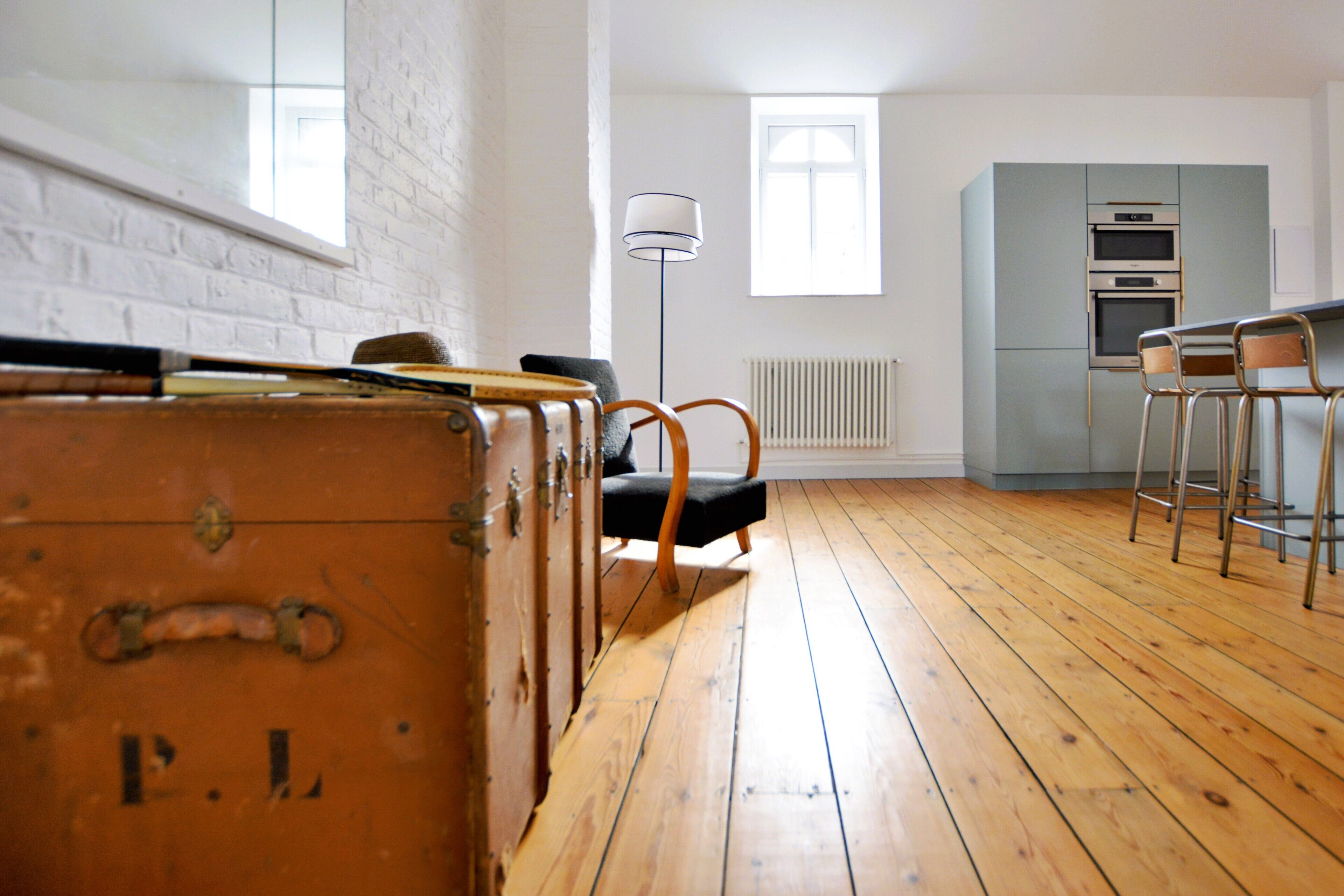 A room with wooden flooring, a vintage suitcase, a black chair, a bar stool, a floor lamp, and a kitchen area with an oven.