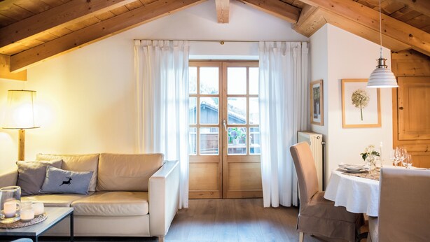 A cozy room with a wooden ceiling, a sofa with cushions, a dining table set for two, and a large window with curtains. - Haus Lesch - Stilvolle Appartements mit tollem Bergblick in Kreuth am Tegernsee (Kreuth)