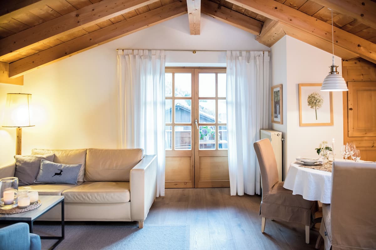 Ein gemütliches Zimmer mit Holzbalkendecke, einer Couch mit Kissen, einem Esstisch für zwei Personen und einem großen Fenster mit Vorhängen.