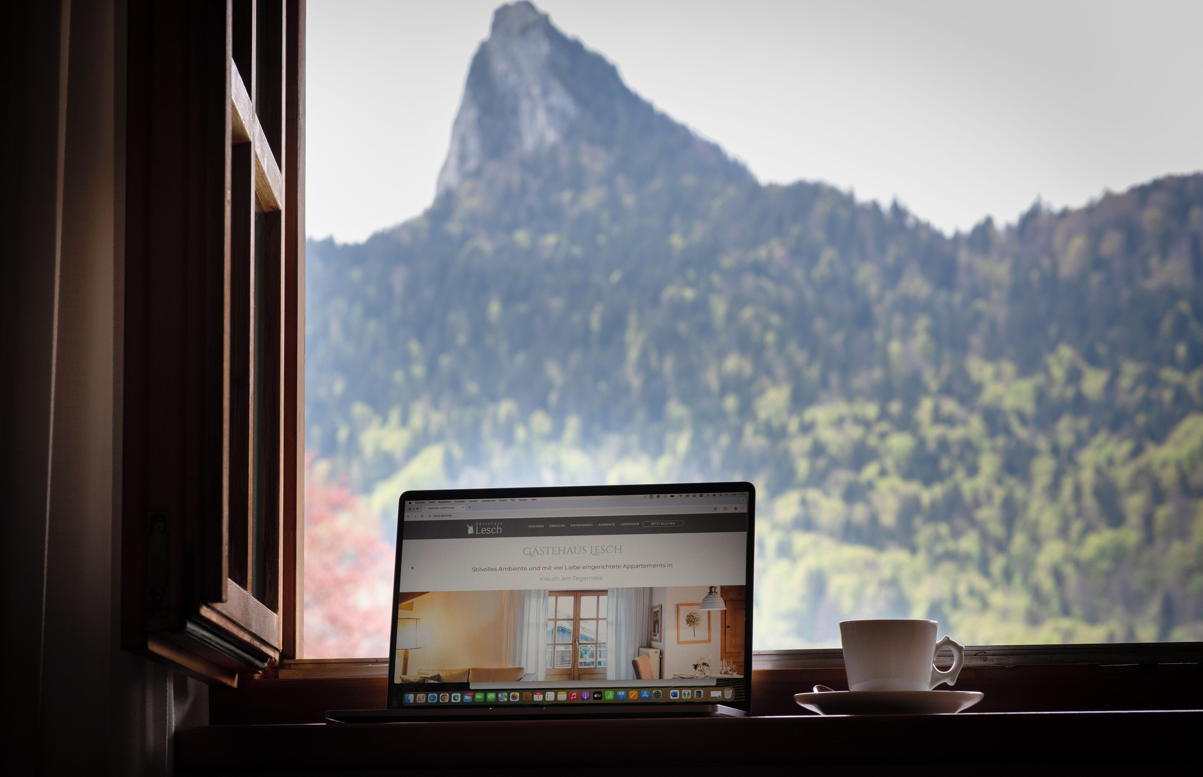 A laptop on a windowsill with a view of a mountain and a webpage titled "Gastehaus LESCH" visible on the screen.