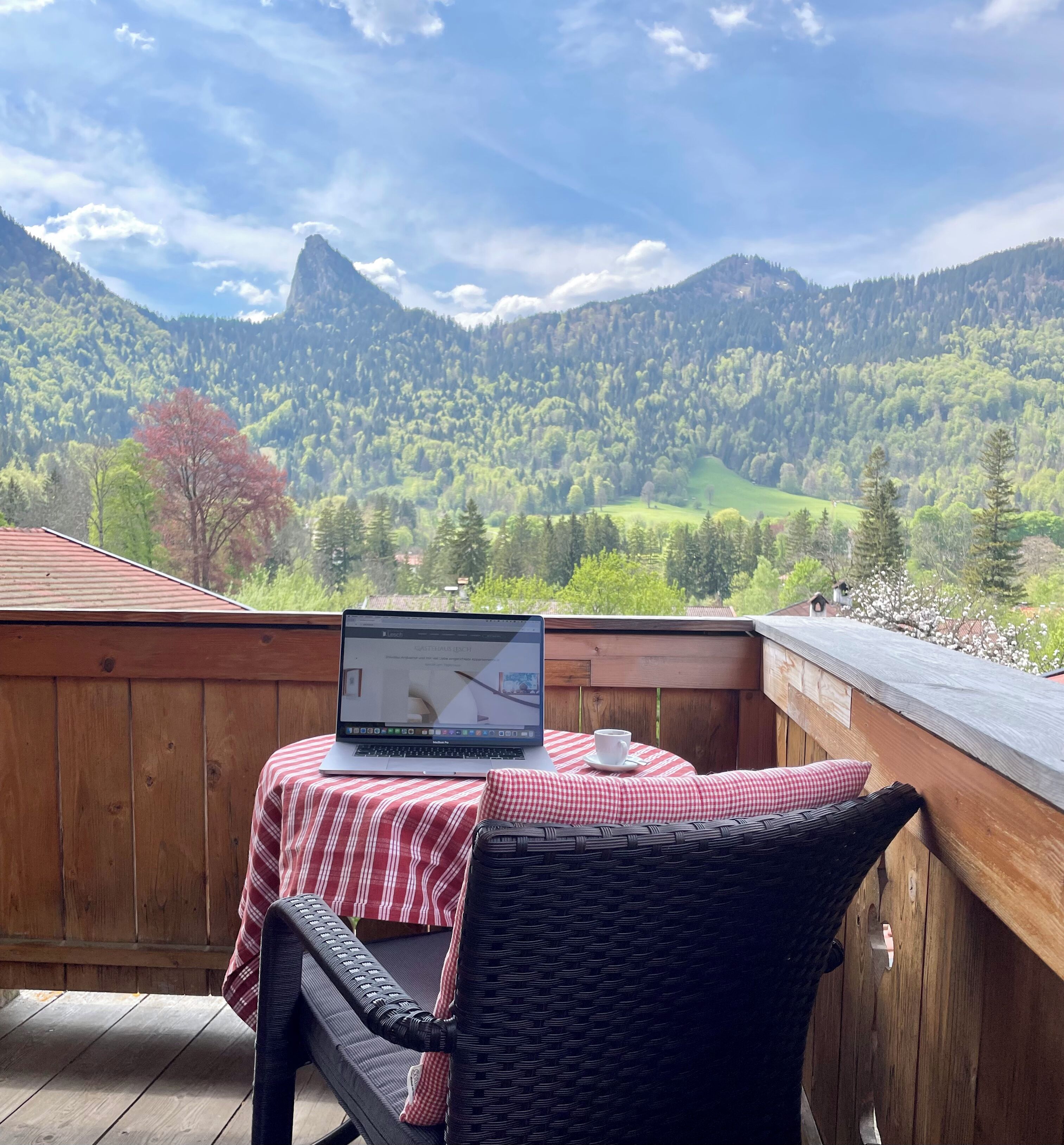 Una terraza de madera con una computadora portátil sobre una mesa, una silla de mimbre y un mantel a cuadros rojo y blanco.