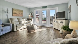 A spacious living room with a beige sofa, a patterned armchair, a television, and a large window with a view of a balcony. - Direct ocean view Villa Capriani - just renovated! (North Topsail Beach)