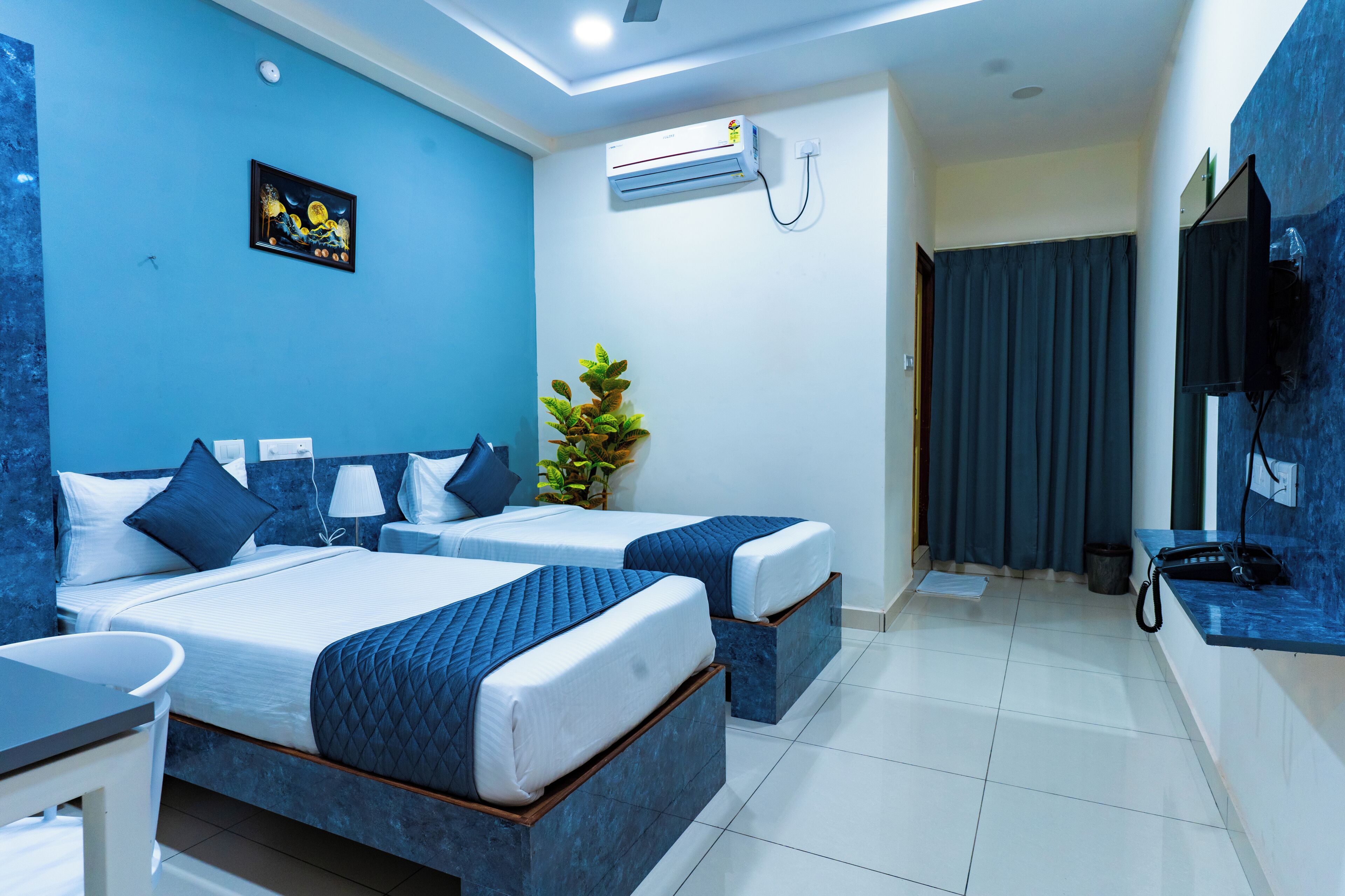 Une chambre d’hôtel avec deux lits, un bureau, une chaise, une plante, un climatiseur et un téléviseur fixé au mur.