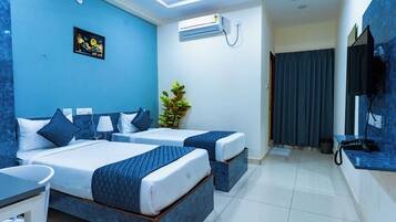 A hotel room with two beds, a desk, a chair, a plant, an air conditioning unit, and a TV mounted on the wall.