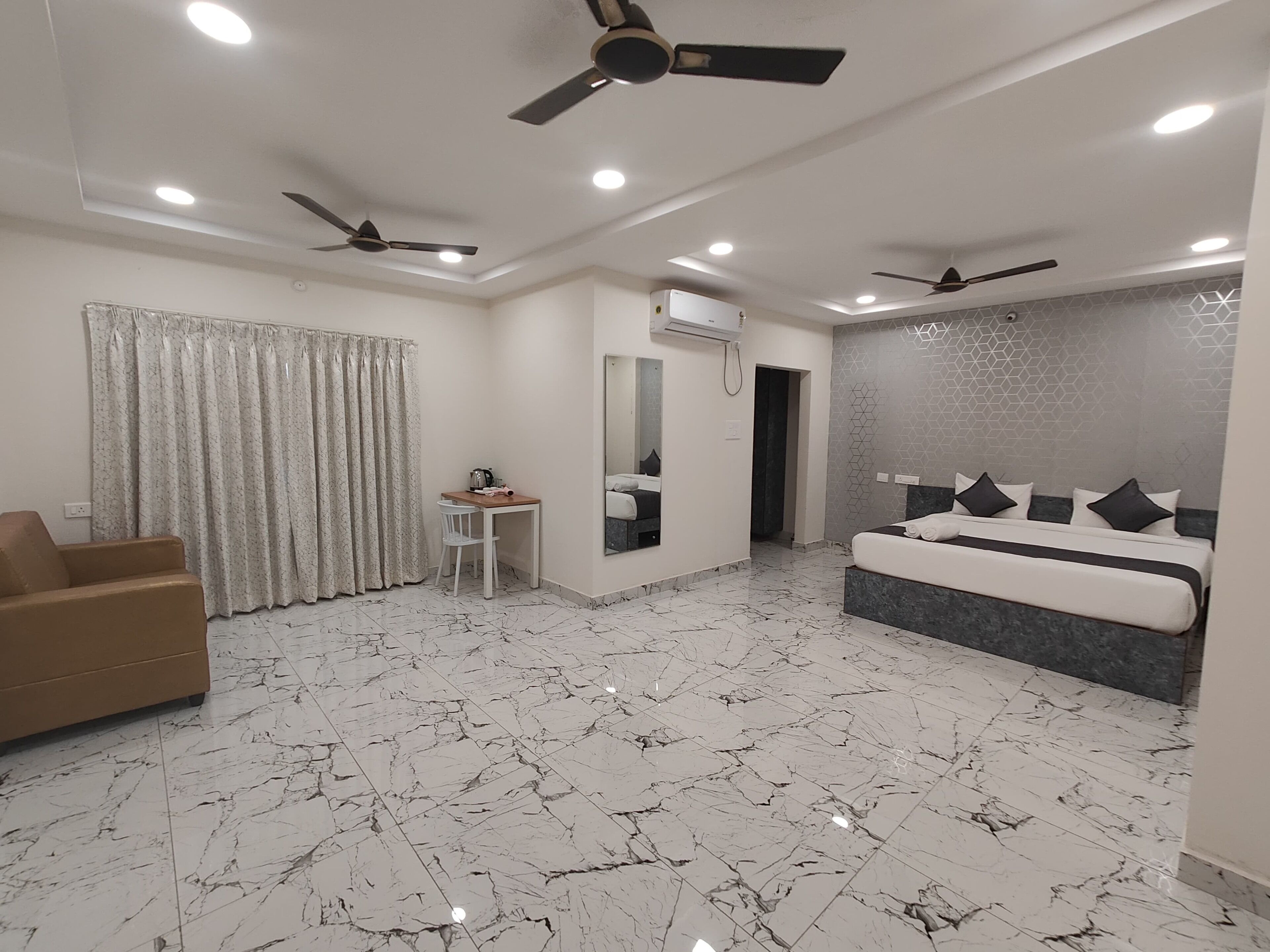 Une chambre d’hôtel moderne avec un sol en marbre, un lit avec une literie noire et blanche, un fauteuil marron, une petite table avec une bouilloire et deux ventilateurs de plafond.