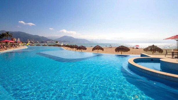 A clear blue pool with a curved edge, surrounded by beach umbrellas and palm trees, with mountains in the background. - Grand Venetian Beachfront Condo (Puerto Vallarta)