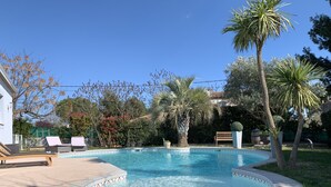 A swimming pool with a tiled edge, surrounded by palm trees and other greenery. There are lounge chairs and a bench near the pool. - Belle Villa Familiale Tout Confort à 15 mn des Plages (Cournonterral)