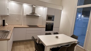 A modern kitchen with white cabinetry, a built-in oven, microwave, and a gas stove. There is a dining table with four chairs and a bowl on it. The floor is wooden, and the ceiling has a unique curved design.
