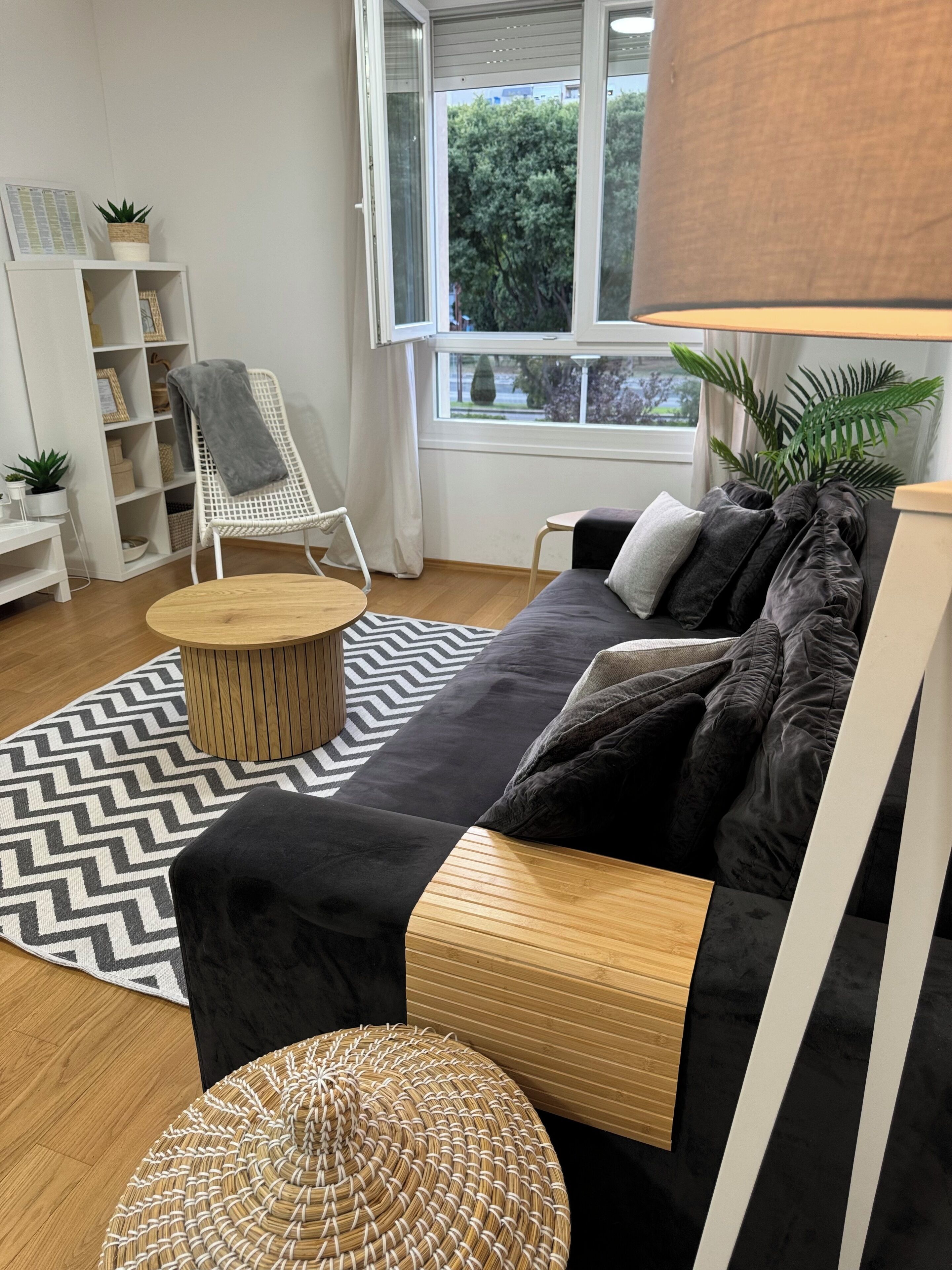 Ein modernes Wohnzimmer mit einer schwarzen Couch, einem Holztisch, einem gewebten Hocker, einem weißen Sessel und einem Bücherregal.