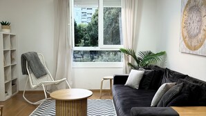 A modern living room with a black sofa, a round wooden coffee table, a white chair, and a view of a cityscape through a large window. - Allora (Split)