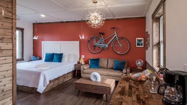 A modern room with a wooden floor, a bed with white bedding, a sofa with blue cushions, a wooden coffee table, a bicycle mounted on the wall, and a coffee machine. - Extended Stay | With Kitchen | Prescott Studio (PRESCOTT)
