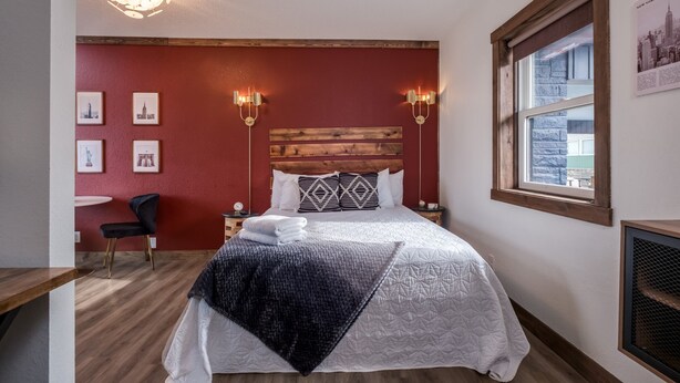 A bedroom with a wooden headboard, a bed with patterned bedding, two wall-mounted lamps, and a window with a view of buildings. - Work & Stay | Free Parking | Simple Comfort (Prescott)