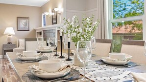 Un coin repas avec une table dressée pour quatre personnes, comprenant des assiettes, des verres et des couverts. Un vase avec des fleurs blanches est posé sur la table, et la pièce est décorée dans des tons neutres, avec des murs de couleur beige et une grande fenêtre.