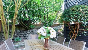 Une table en bois, des chaises en osier, un vase avec des fleurs blanches et, au fond, un mur de pierre.
