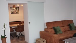 Ein gemütlicher Wohnbereich mit braunem Sofa, grünen Kissen, einem Esstisch mit Stühlen und einem Bild an der Wand.
