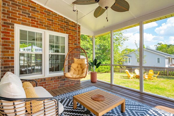A covered porch with a brick wall, a hanging chair, a wooden table, and a view of the backyard. - Hanahan Haven GameRoom Bikes FirePit Dog & Family Friendly (Hanahan)