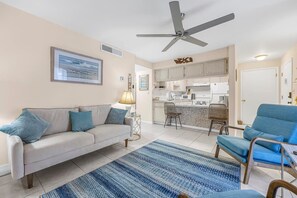 Un salon avec un canapé, un fauteuil bleu, un tapis à motifs, un ventilateur de plafond et, au fond, une cuisine.