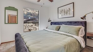 Une chambre à coucher avec un lit, un ventilateur de plafond, une fenêtre donnant sur des arbres et un tableau encadré au mur.