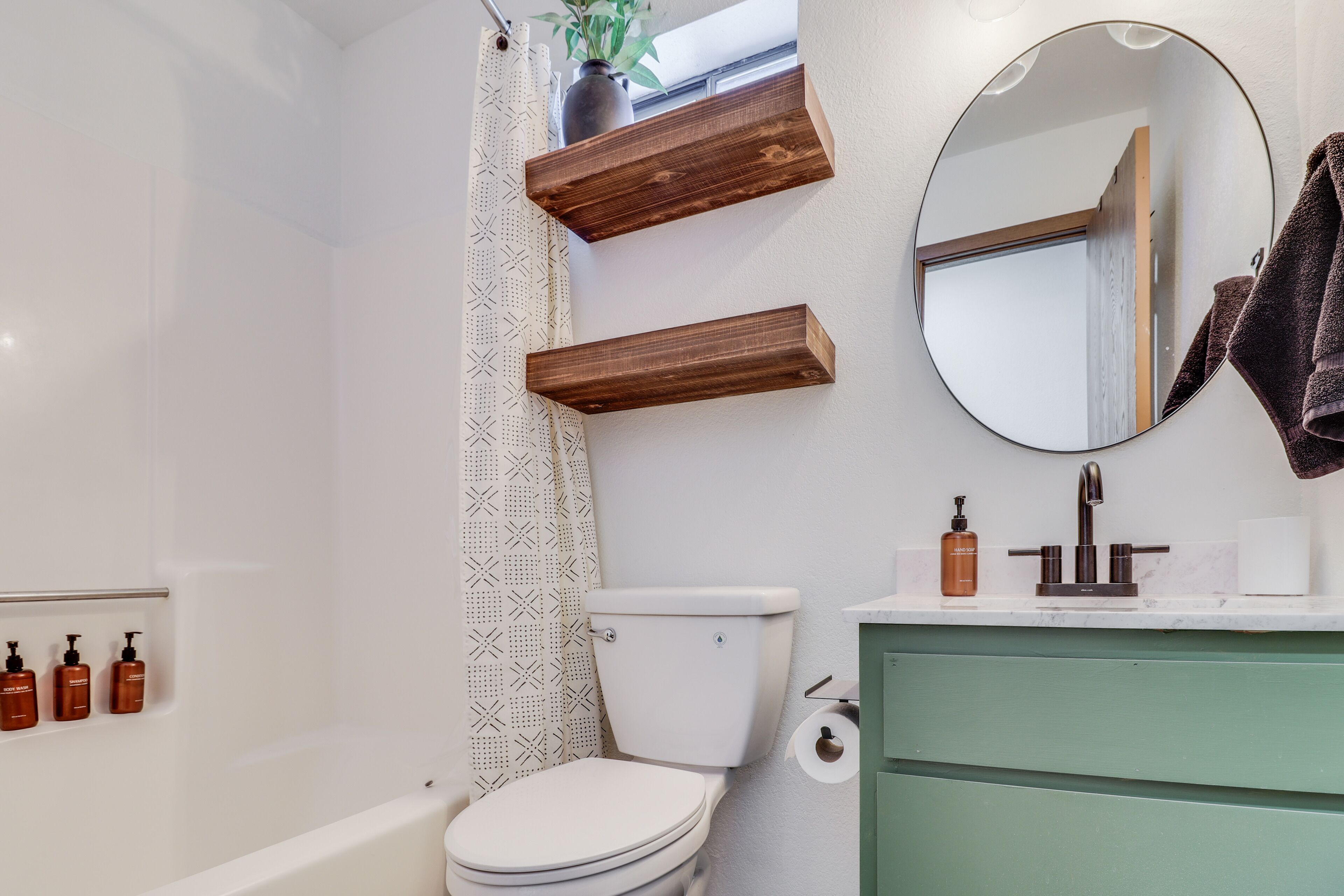 A bathroom with a white toilet, a green vanity, wooden shelves, a round mirror, and a shower curtain with a geometric pattern.