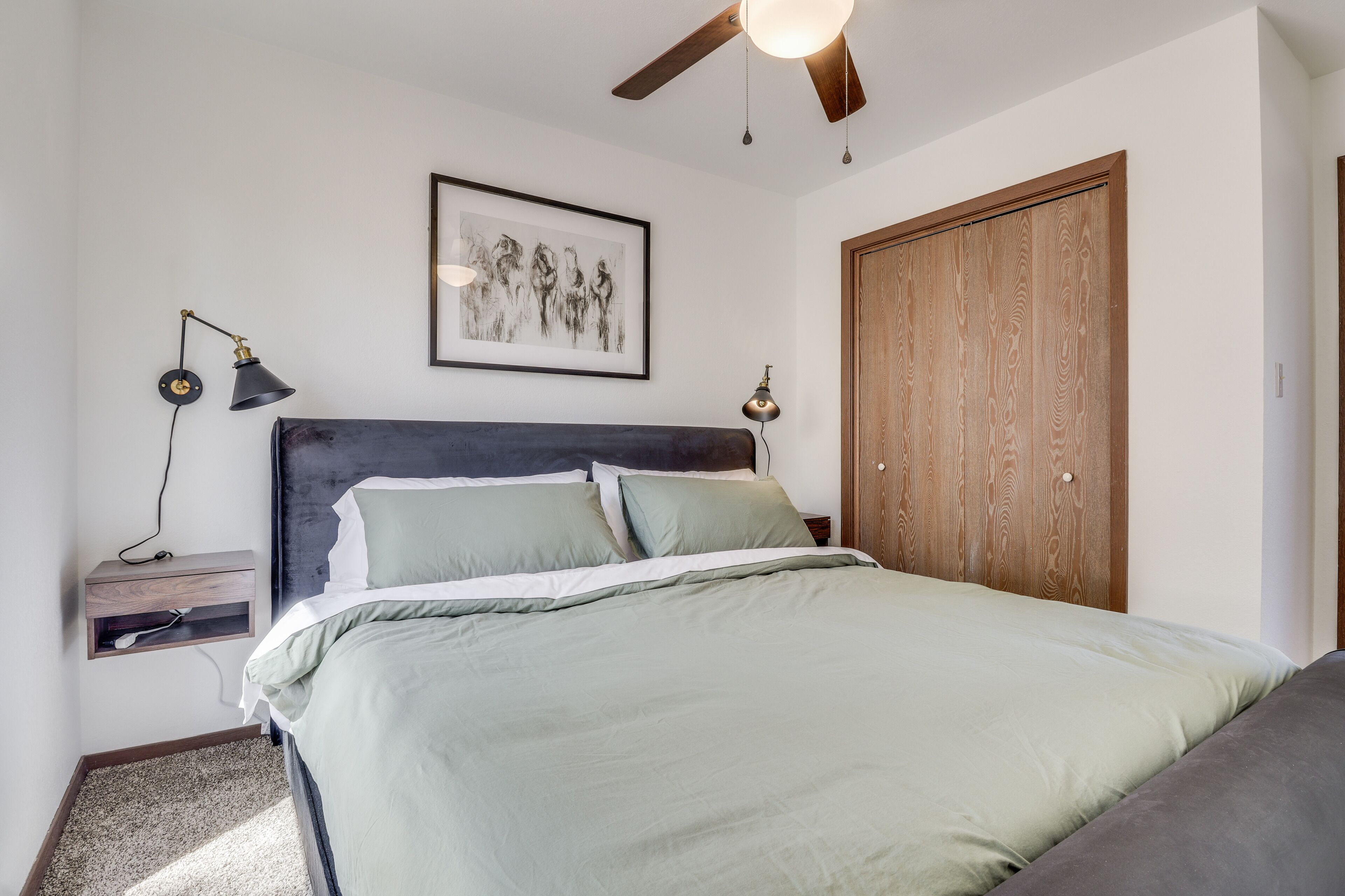 Ein Schlafzimmer mit Bett, Nachttisch, Schreibtisch, einem gerahmten Bild, einem Holzschrank und einem Deckenventilator.