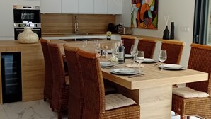 Une cuisine moderne avec des meubles blancs, un four encastré et un coin repas doté d’un sol en marbre et de chaises en osier.