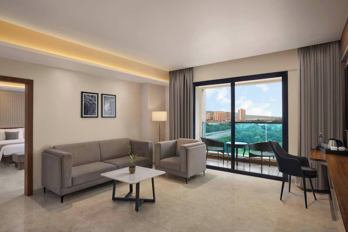 A modern hotel room with a sofa, a small table, a lamp, and a view of the city through large windows.