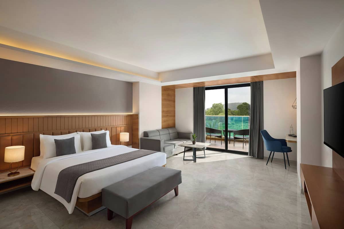 A modern hotel room with a large bed, a desk, a sofa, and a balcony with a view.