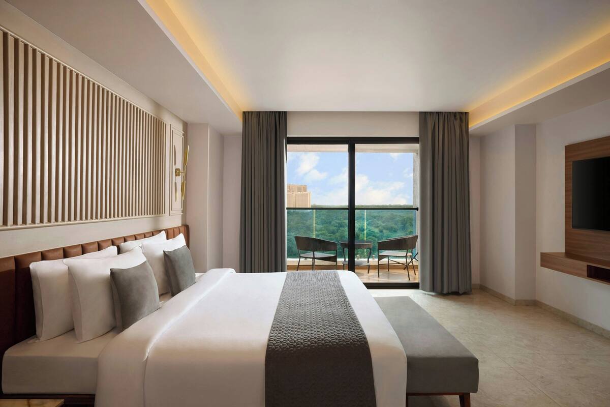 A hotel room with a large bed, a TV mounted on the wall, and a balcony with chairs.