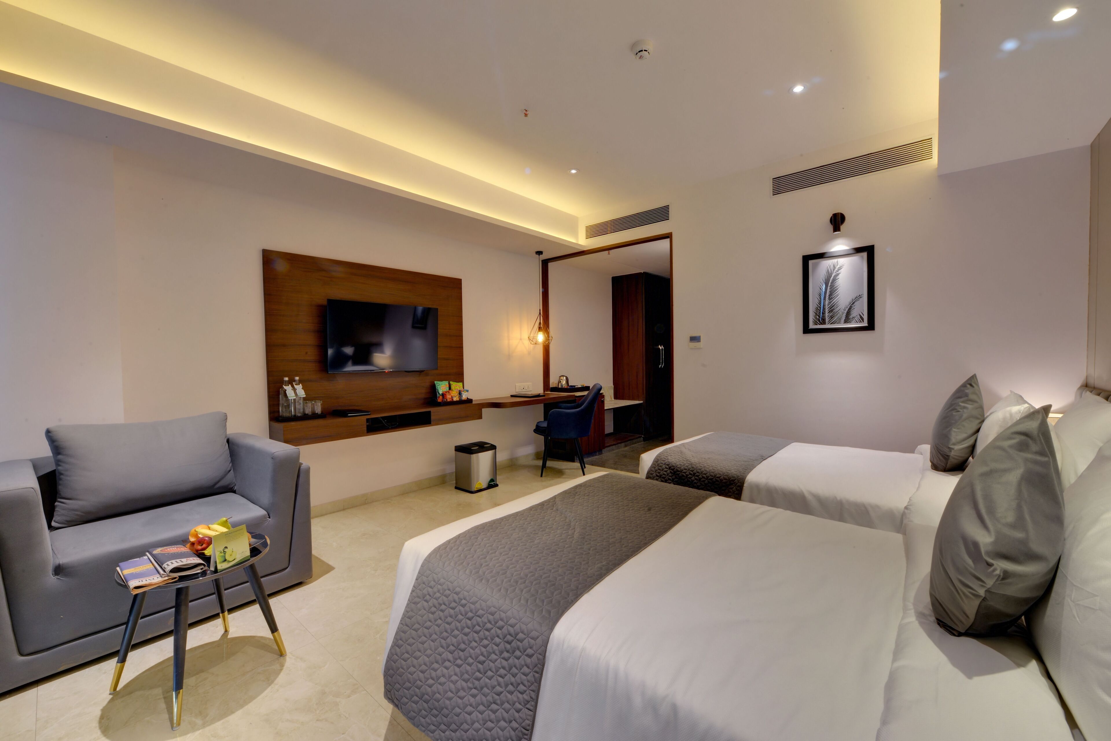 A modern hotel room with a sofa, two beds, a desk, and a TV mounted on the wall.
