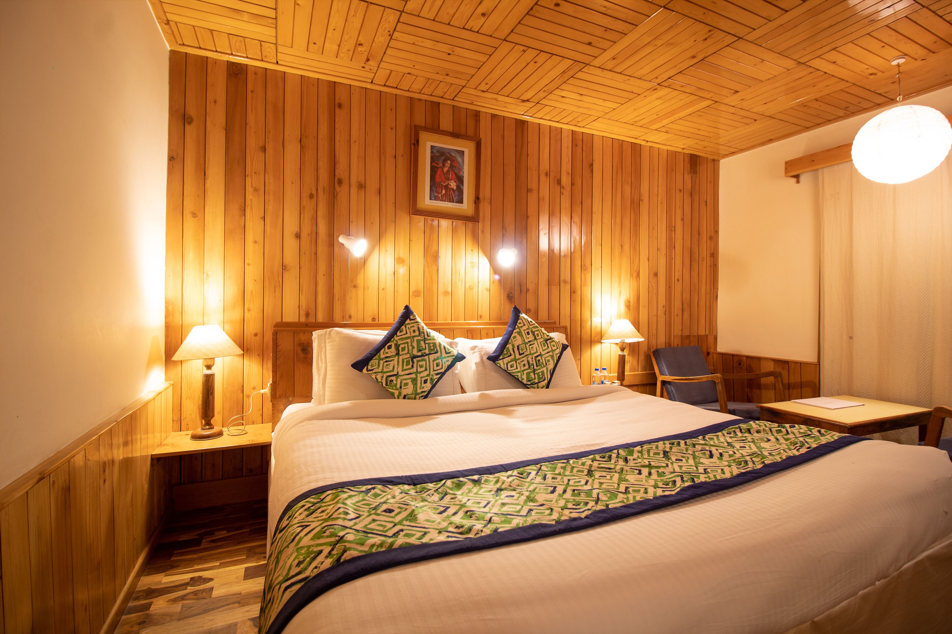 A wooden-paneled room with a bed, two bedside tables with lamps, a chair, and a desk with a chair.