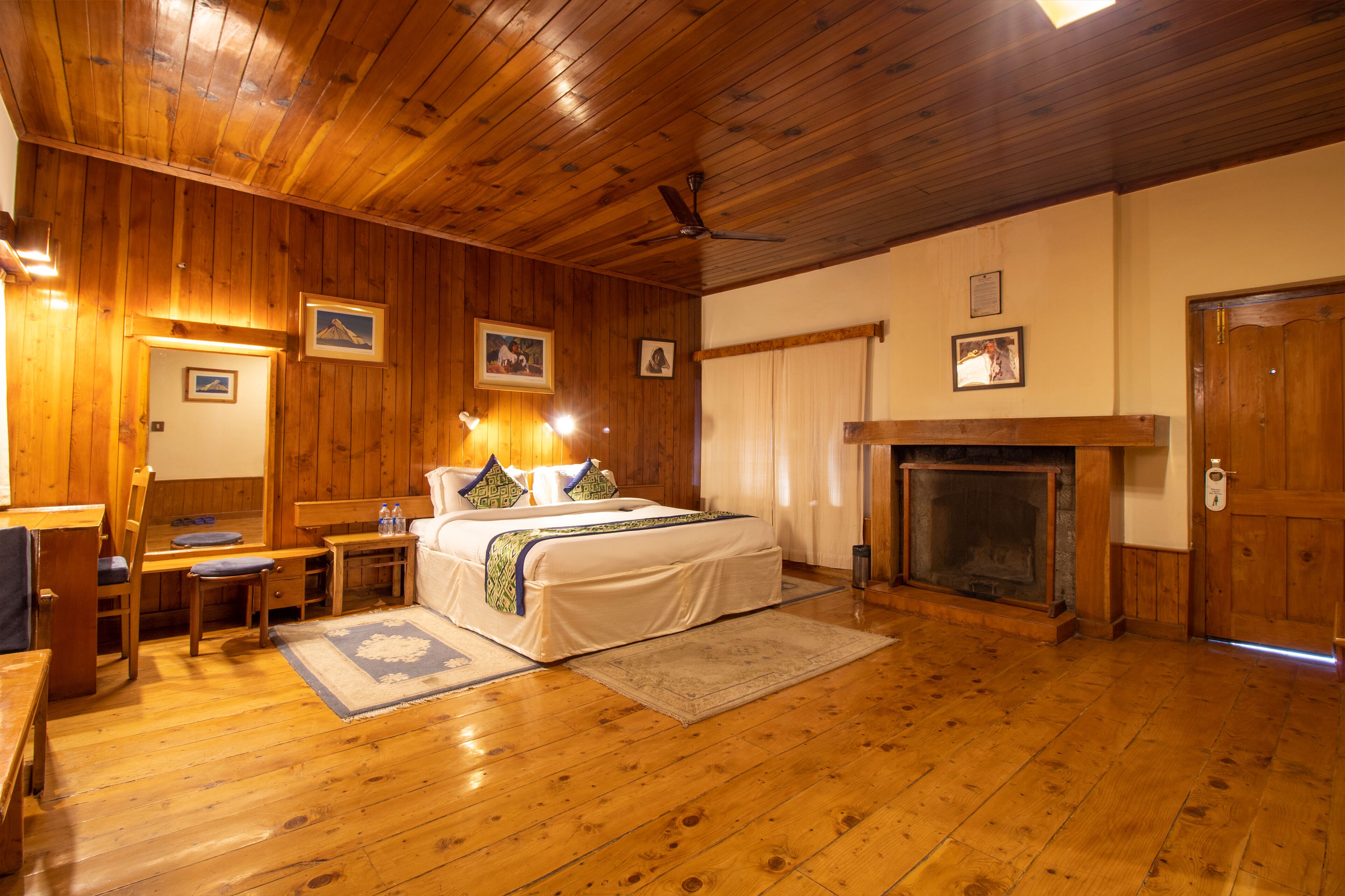 A wooden-floored bedroom with a bed, bedside tables, a fireplace, and framed pictures on the walls.