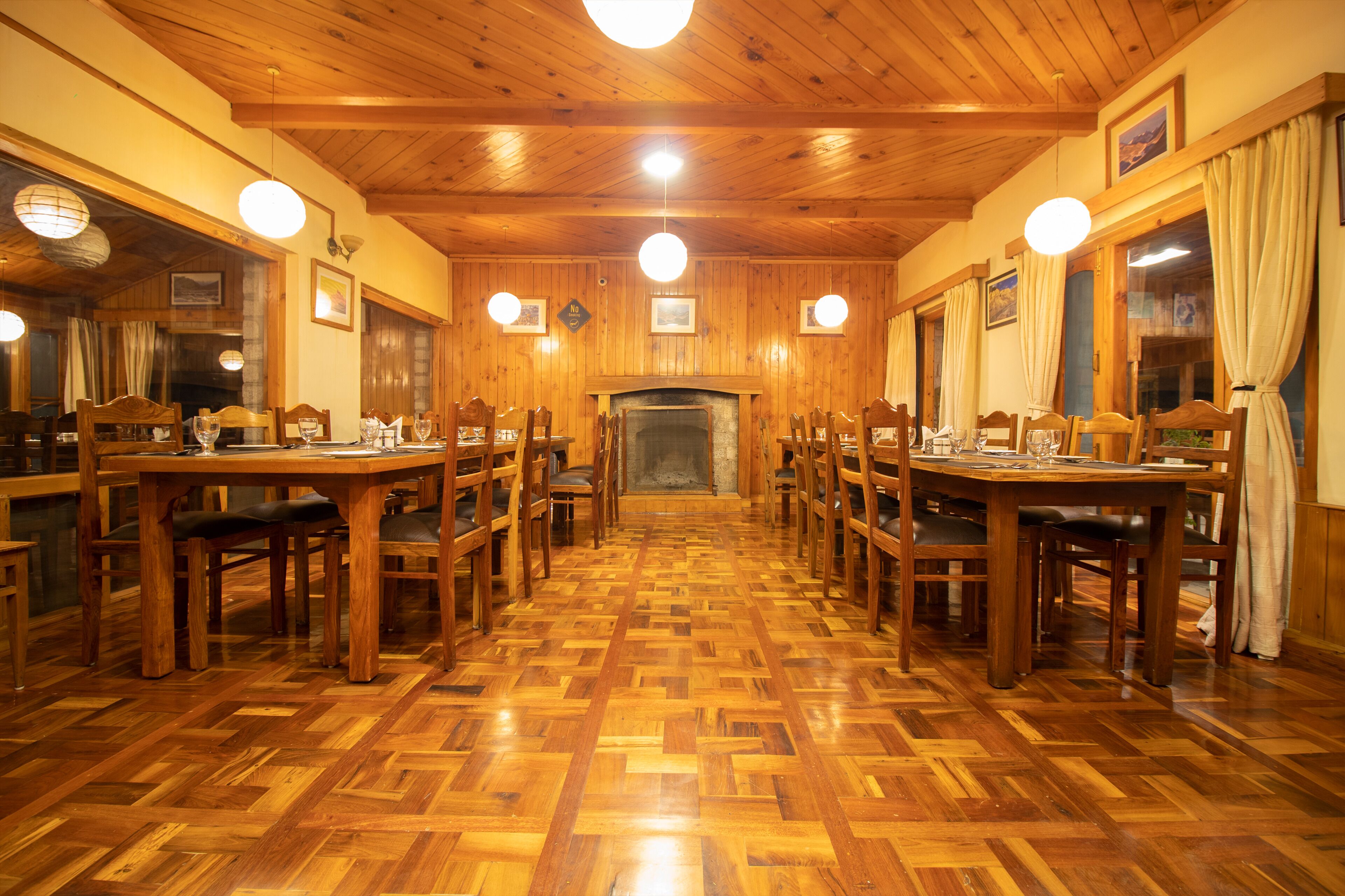 Un espace repas avec des tables et des chaises en bois, une cheminée et de grandes fenêtres dotées de rideaux.